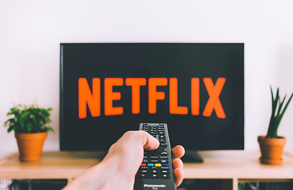 En hand hanterar sakkunnigt en fjärrkontroll och riktar den mot en TV där Netflix-logotypen lyser. I närheten ger två krukväxter en touch av natur till den eleganta tekniken.
