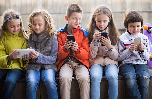 Fem barn sitter på rad med varsin smartphone eller surfplatta, helt uppslukade av tekniken. Klädda i lediga friluftskläder är de vilse i sina digitala världar. En skateboard vilar bredvid dem och antyder det roliga som väntar när skärmtiden är över.